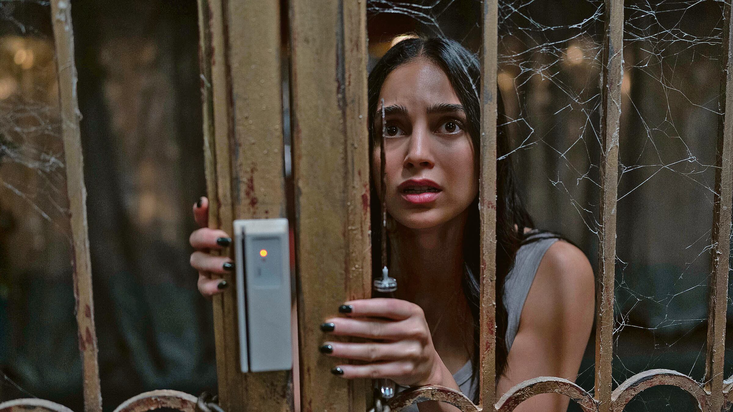Melissa Barrera en personaje, con cara de terror tras un portón de rejas amarrado con cadenas. Sostiene un cuchillo.